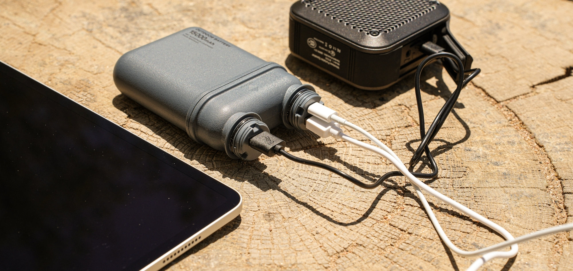 NESTOut 5000 mAh Power Bank in Grey Color charging speaker and iPad Outside on a Tree Stump