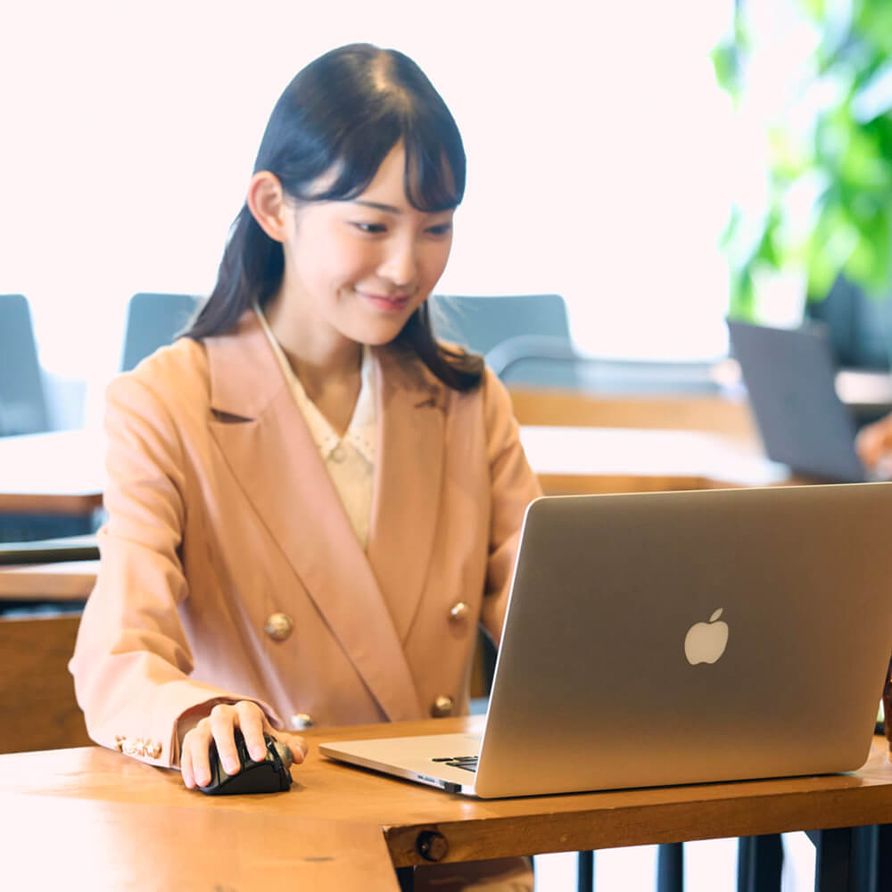 A woman in a peach blazer sits at a wooden desk using a silver Apple laptop and black bitra thumb trackball mouse in a bright, modern office setting.