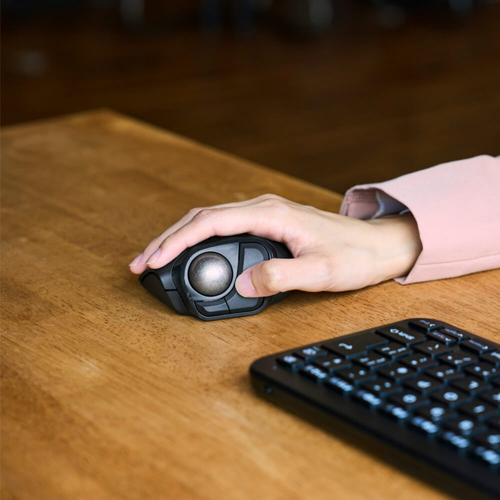 A close-up of a person's hand using a black bitra thumb trackball mouse on a wooden desk next to a keyboard.