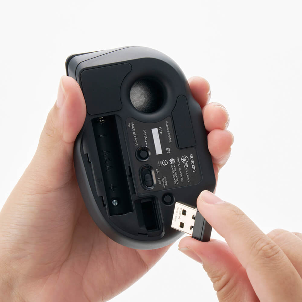 A hand holds the underside of a black bitra index trackball mouse, showing battery compartments and a USB port, while another hand inserts a USB flash drive.