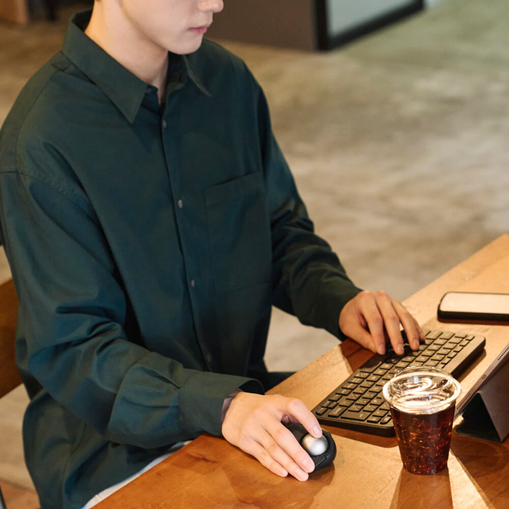 A person wearing a dark green long-sleeve button-up shirt sits at a wooden desk using a computer bitra index trackball mouse and keyboard, with a coffee cup nearby.