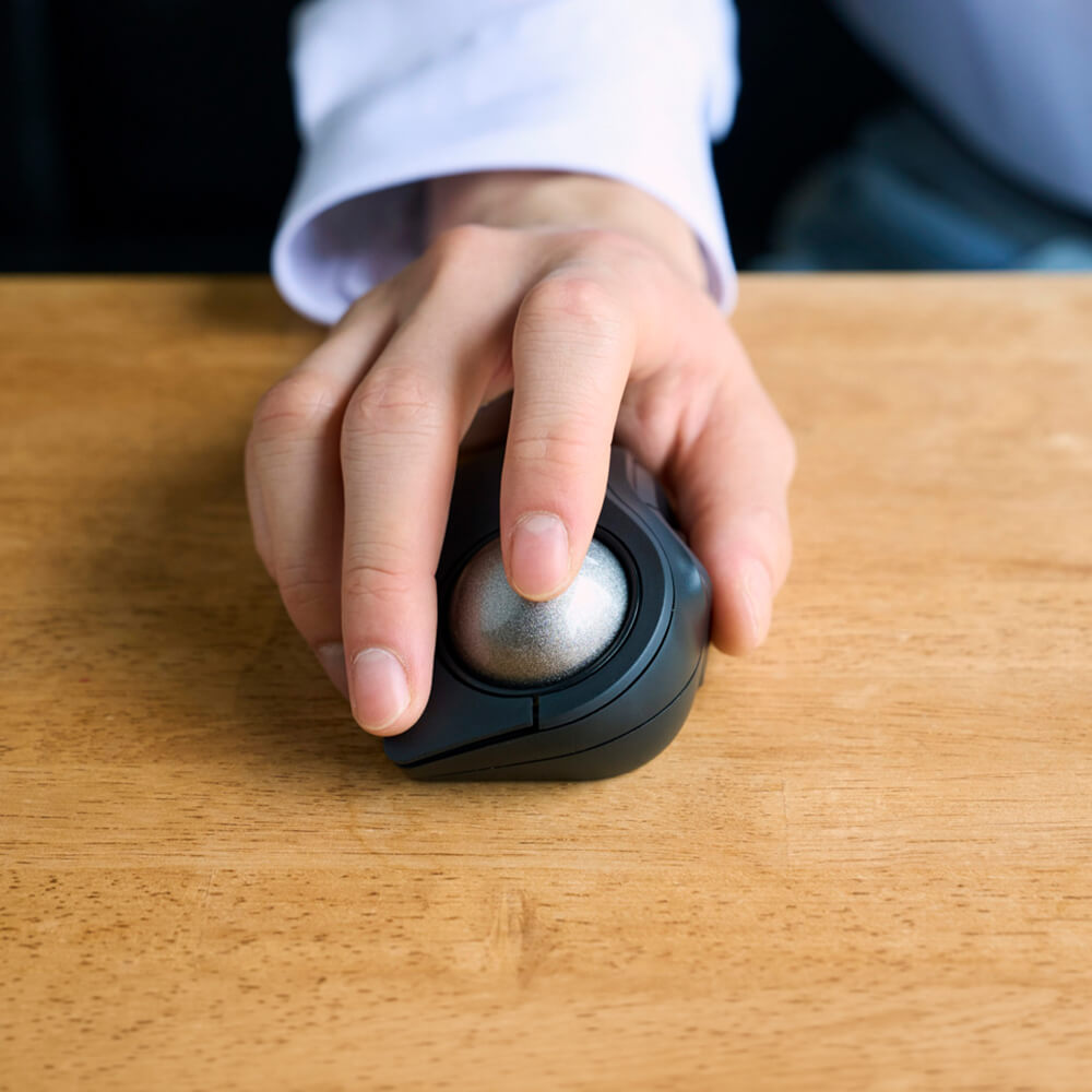 A close-up of a person's hand using a black bitra index trackball mouse on a light brown wooden desk.
