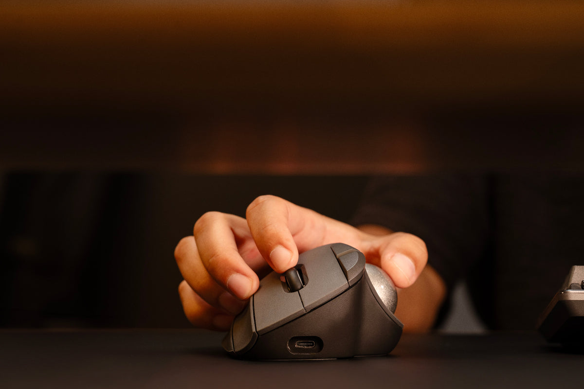 A close-up of a hand using a black wireless computer mouse on a dark desk surface, with a warm blurred background.