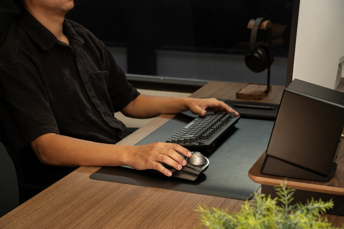 A person in a black shirt uses a sleek black trackpad with a circular silver touchpad on a wooden desk, alongside a keyboard, monitor stand, and headphones.