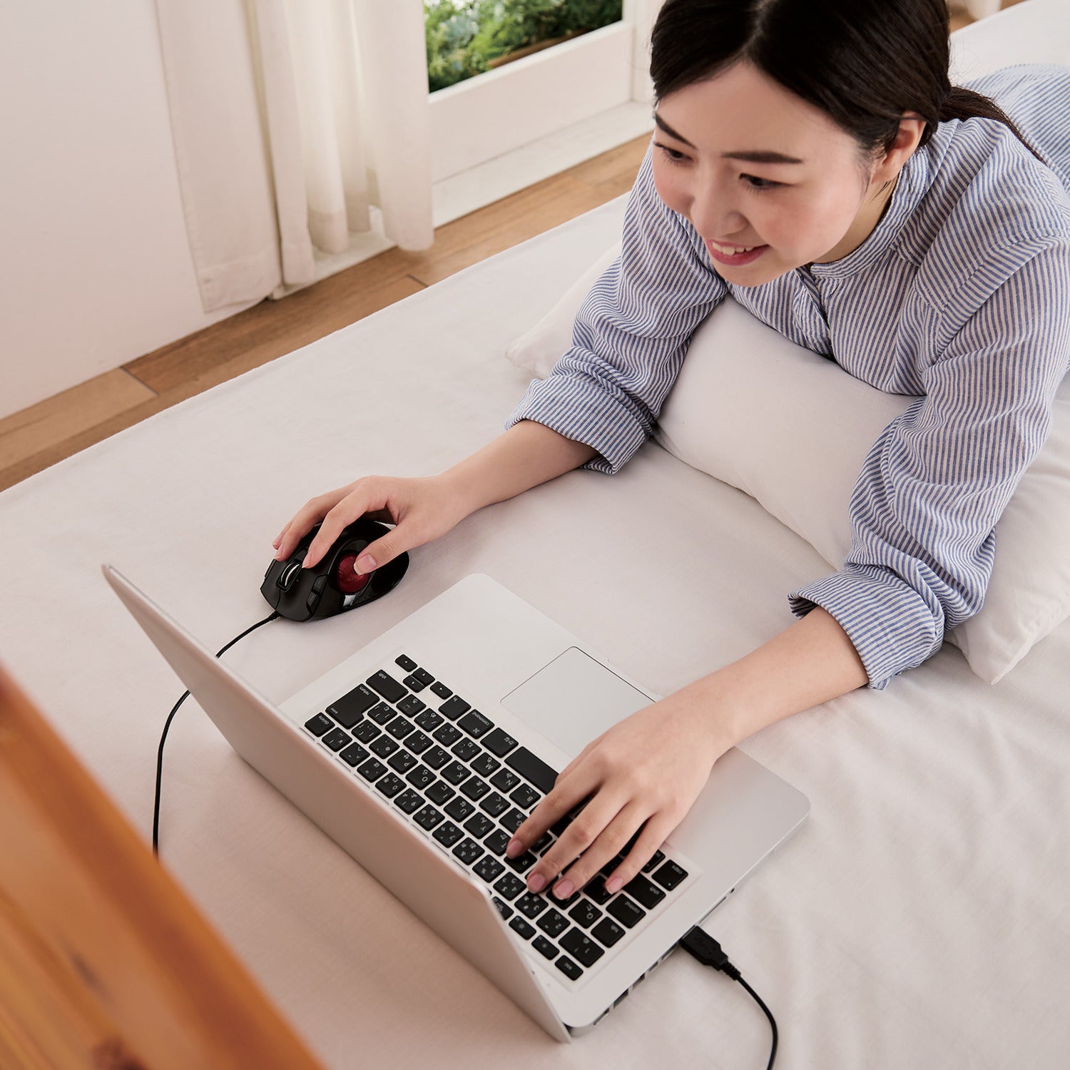 Woman laying stomach down on bed using Black EX-G Trackball Wired Red Ball connected to laptop | Elecom Wireless Trackball mouse