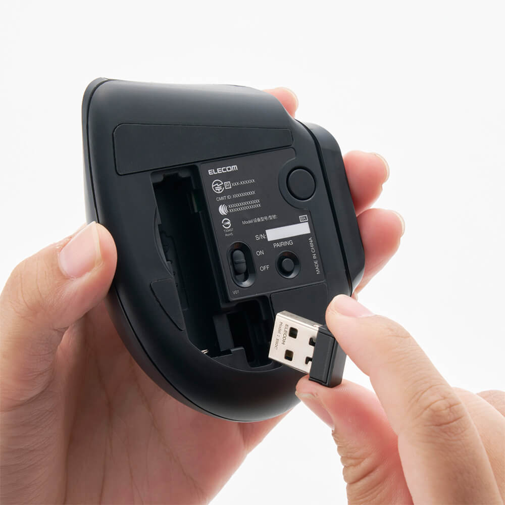A person's hands holding a black Elecom wireless bitra thumb trackball mouse underside to show the open battery compartment and USB port, with a USB receiver being plugged in against a white background.