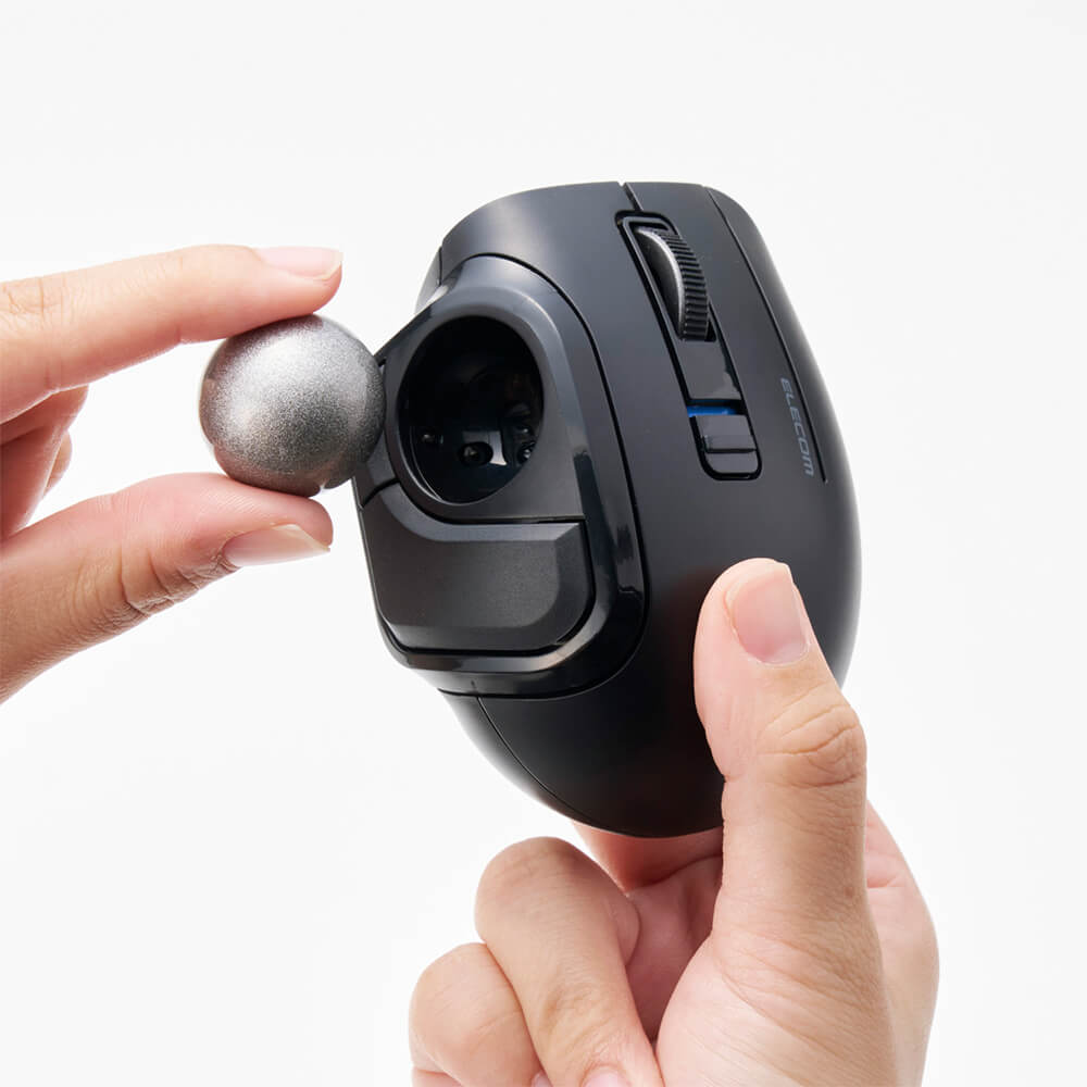 A black ergonomic bitra thumb trackball mouse being held by two hands, with a small metallic silver ball resting against its side. The background is a clean, solid white.