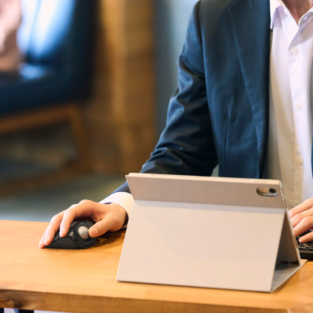 A close-up of a person in a navy blazer and white shirt using a tablet in landscape mode on a wooden desk, with a black bitra thumb trackball mouse in their other hand.