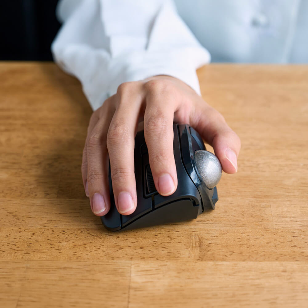 A close-up of a hand using a black bitra thumb trackball mouse with a silver thumb wheel on a wooden desk, with a person's white shirt visible in the background.