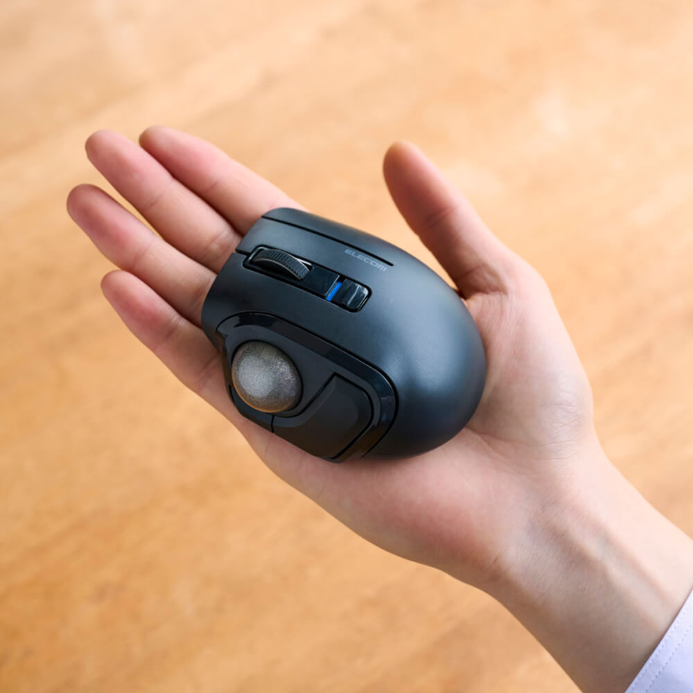 A ergonomic black bitra thumb trackball mouse with a thumbwheel and blue accent buttons, held in a person's hand against a warm wooden background.