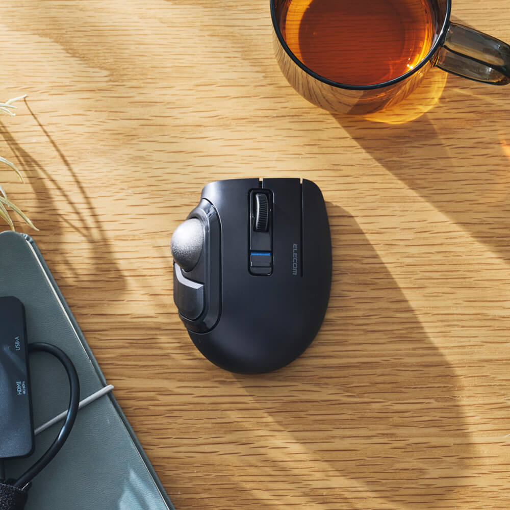 A black ergonomic bitra trackball mouse with a thumbwheel and blue scroll accent, placed on a wooden desk next to a glass mug of tea and a notebook.