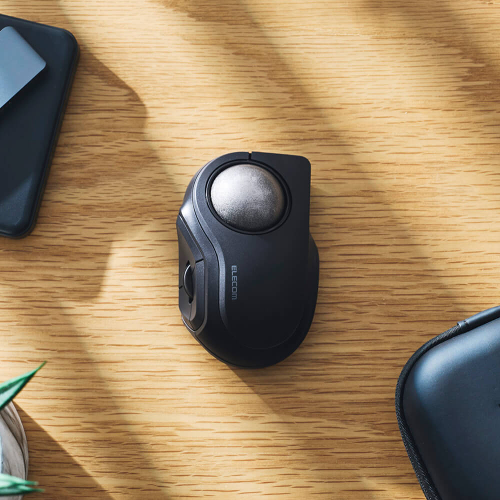 A black bitra index trackball mouse with a circular top, placed on a light wood desk with soft shadows.