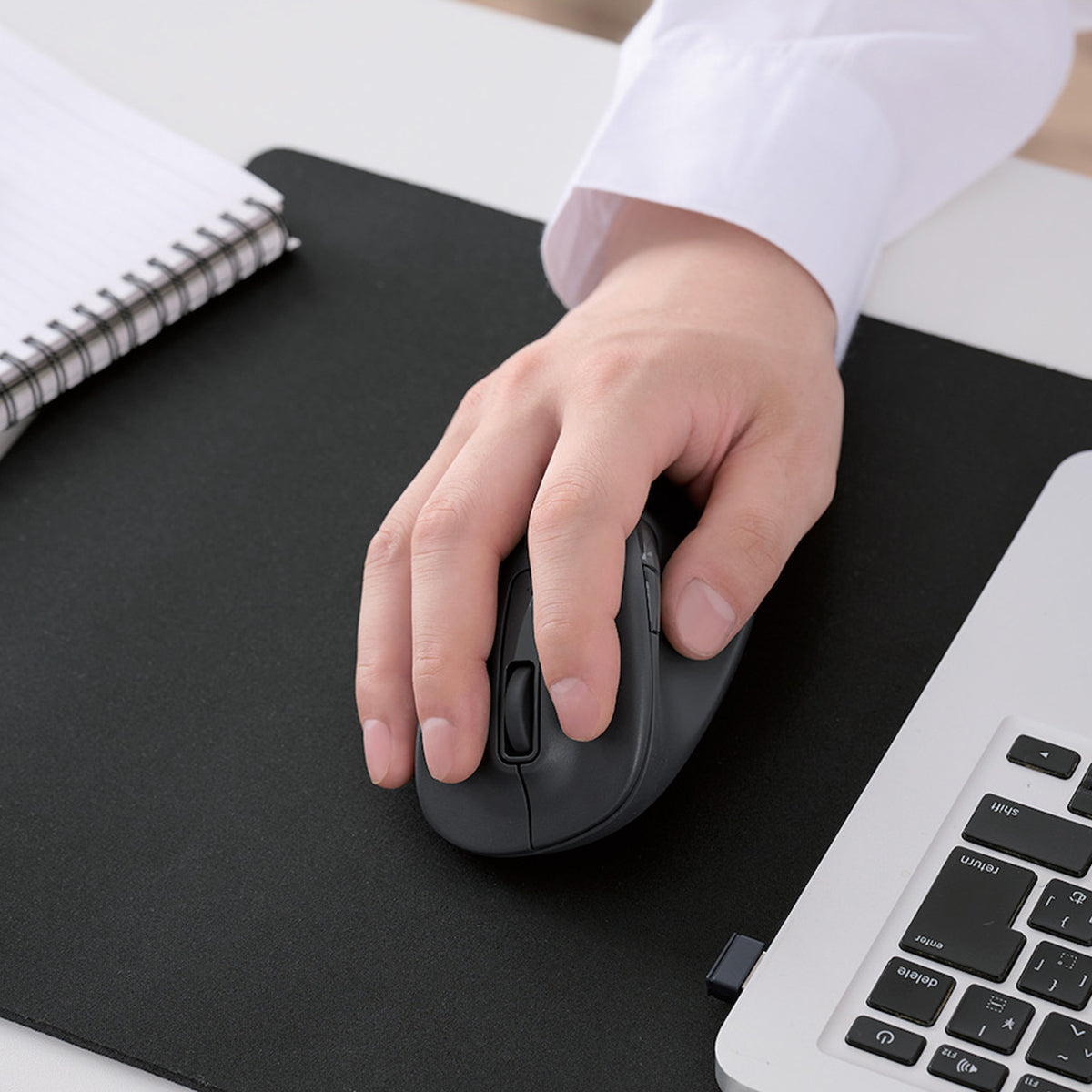 A person's hand in a white shirt using a black wireless computer mouse on a large black desk mat, with a laptop keyboard and spiral notebook visible.